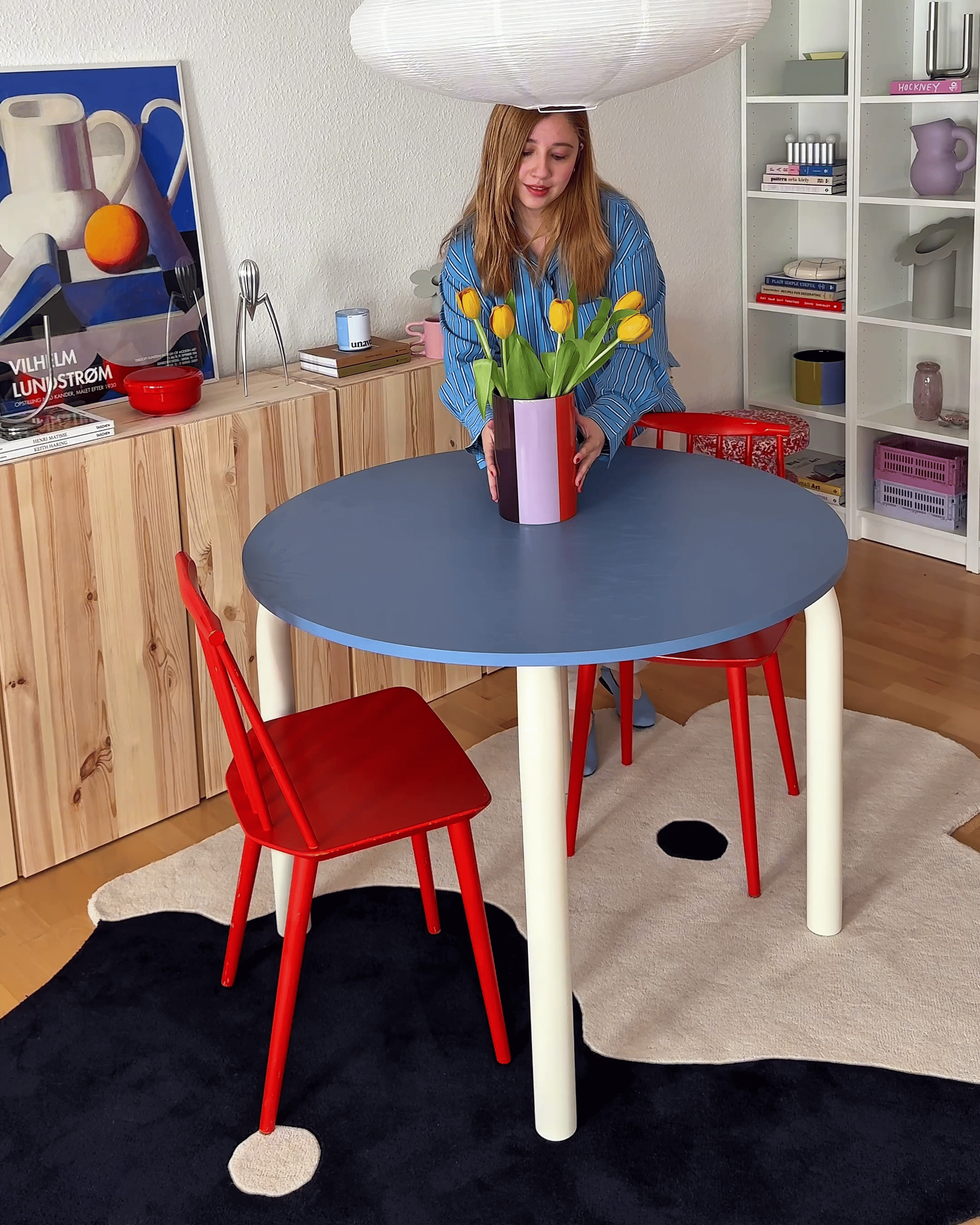 Woman arranging tulips in a vase at a round blue table with red chairs in a room.