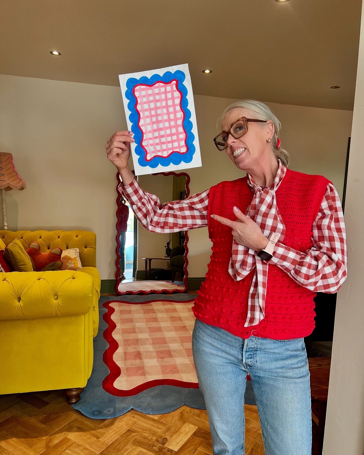 Person in a red checkered shirt and red vest holding up a colorful card in a living room.